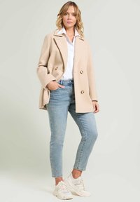 Abrigo beige de corte doble con botones dorados, combinado con jeans de lavado claro y zapatillas blancas; tela suave, diseño clásico, silueta estructurada.