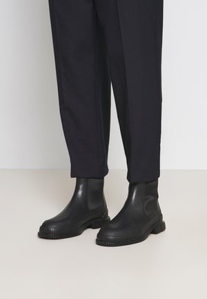Botas negras hasta el tobillo con paneles laterales elásticos, suelas de goma y detalles texturizados. Combinadas con pantalones oscuros a medida frente a un fondo neutro.