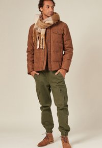 Brun quiltad jacka med knappar, gröna cargobyxor och bruna mockaankelstövlar. Själen har beiga och gyllene nyanser med fransiga kanter.