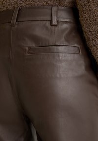 Bruine leren broek met een gladde textuur, voorzien van een enkele achterzak en subtiele stiksels, gepresenteerd tegen een pluizige stofachtergrond.