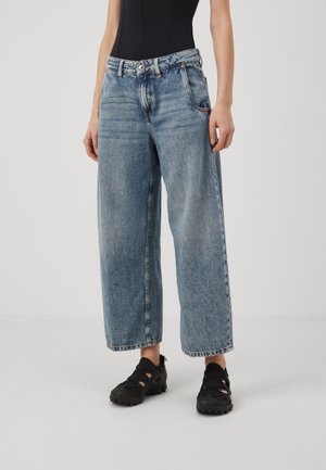 Personne portant un jean ample bleu clair à jambes larges, associé à des sandales noires avec des découpes, debout devant un fond uni.