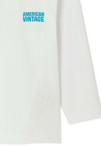 Chemise blanche à manches longues avec le texte bleu "AMERICAN VINTAGE" imprimé sur la partie supérieure gauche de la poitrine.