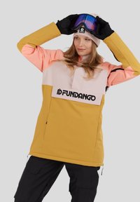 Woman adjusting blue ski goggles wearing a color-blocked yellow, pink, and beige jacket and black gloves against a gray background.