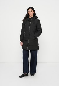 Svart polstret jakke med glidelås, hette og to frontlommer. Designet har horisontale quiltede seksjoner og en knelengde.