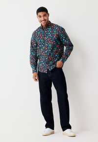 Camicia a maniche lunghe con bottoni, base blu navy e motivi colorati di alberi, attrezzi e forme; abbinata a jeans scuri e sneakers bianche.