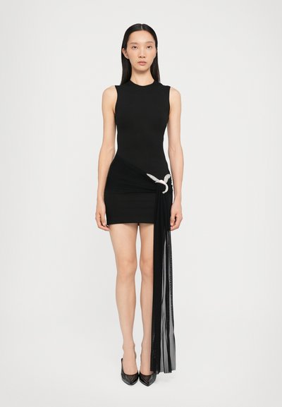 Vestido negro sin mangas con un cuerpo ajustado y una falda drapeada asimétrica con un panel negro transparente, adornado con un detalle plateado.