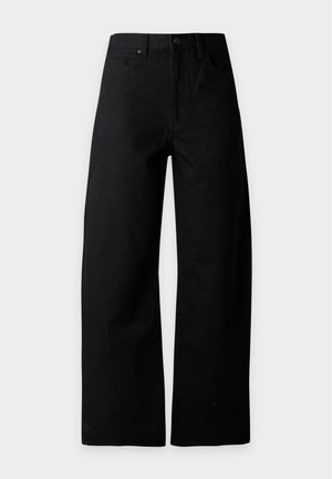 Femme aux longs cheveux noirs portant un pull gris par-dessus une chemise blanche à col, un pantalon noir à jambes larges et des chaussures noires pointues, debout sur un fond blanc.