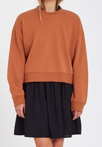 Sweat-shirt marron court avec épaules tombantes, poignets côtelés, et petit logo, associé à une jupe plissée noire. Tenue décontractée en superposition.