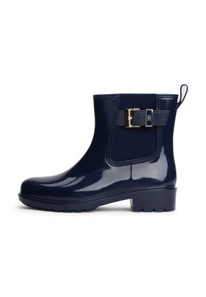 Botte de pluie brillante bleu marine à la cheville avec panneau élastique latéral, sangle à boucle dorée et languette de tirage sur fond blanc.
