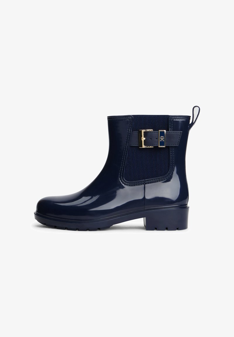 Botte de pluie brillante bleu marine à la cheville avec panneau élastique latéral, sangle à boucle dorée et languette de tirage sur fond blanc.
