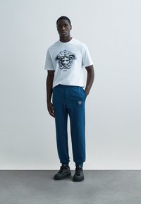 Λευκό βαμβακερό t-shirt με μπλε γραφικό τύπωμα και την επωνυμία "Versace", σε συνδυασμό με μπλε παντελόνι με κεντημένο λογότυπο και μαύρα αθλητικά παπούτσια.
