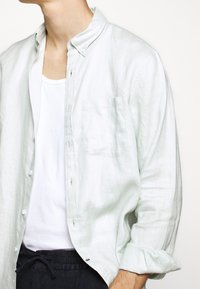 Ljusgrön linneskjorta med knapp-down krage, en ficka och uppvullade ärmar, lagd över en vit linnetopp.