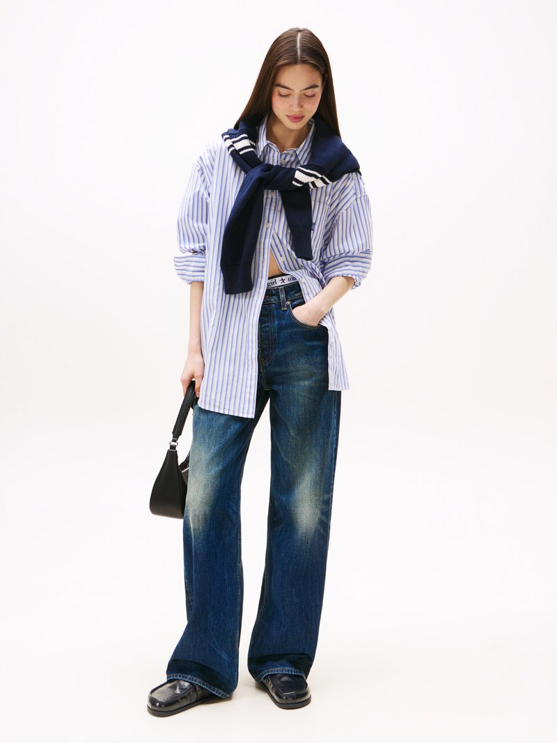 Camicia oversize a righe blu e bianche, jeans blu larghi con una texture sbiadita, maglione blu navy annodato intorno alle spalle, borsa nera, scarpe lucide.