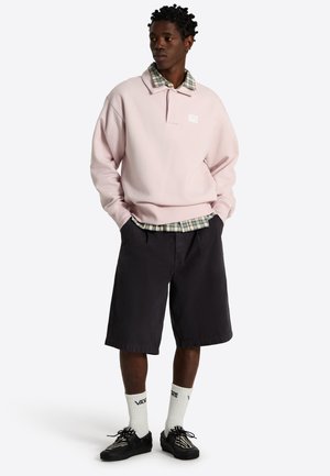 Ung mand iført en lyserød Vans-pullover, ternet skjorte, sorte knælange shorts, hvide Vans-sokker og sorte sneakers, står med hænderne i lommerne.