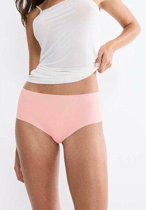 Frau trägt nahtlose Unterwäsche in Hellrosa und ein weißes ärmelloses Oberteil, hebt das Oberteil leicht an, um den Hosenbund zu zeigen.