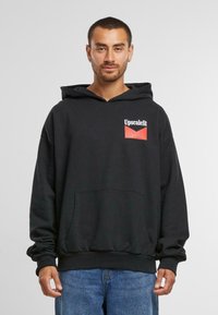 Felpa nera con cappuccio e tasca frontale, caratterizzata da un patch con logo rosso e bianco sul lato sinistro del petto. Tessuto morbido, vestibilità comoda, polsini e orlo a coste.