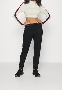 Kobieta w czarnych spodniach dresowych z bocznymi paskami i beżowej krótki rękaw topie, stoi z rękami na biodrach na białym tle.