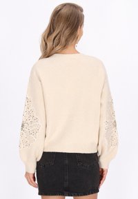 Beige kötött pulóver, virágos díszítéssel az ujjakon, csillogó részletekkel és bordázott aljjal, fekete farmer szoknyával párosítva.