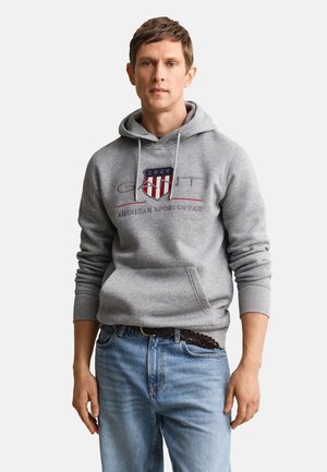 Uomo che indossa una felpa grigia con cappuccio con il logo "1949 GANT American Sportswear" e jeans azzurri chiari, in piedi davanti a uno sfondo bianco uniforme.