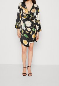 Robe noire à encolure en V profonde, à manches longues, avec un motif floral en crème et jaune, et une taille nouée. Associée à des sandales noires à lanières.