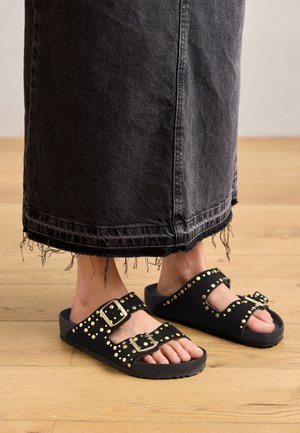 Pieds portant des sandales noires avec des clous dorés et des boucles, associés à une jupe en denim noire à ourlet effiloché, debout sur un sol en bois.