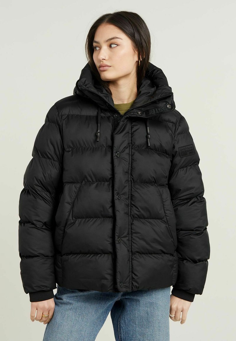 Schwarze Steppjacke mit Kapuze, Knopfverschluss, Kordelzug und gestepptem Muster. Verfügt über einen Aufnäher und gerippte Bündchen. Wird über einem grünen Oberteil getragen.