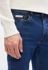 Niebieskie dżinsy Calvin Klein o gładkiej fakturze, z metką na tylnej kieszeni i kontrastowymi szwami, noszone z dzianinowym białym swetrem.