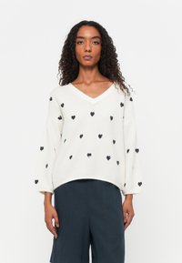 Maglione crema con scollatura a V e maniche oversize, caratterizzato da motivi a cuore neri. Texture morbida con una vestibilità comoda. Abbinato a pantaloni scuri.