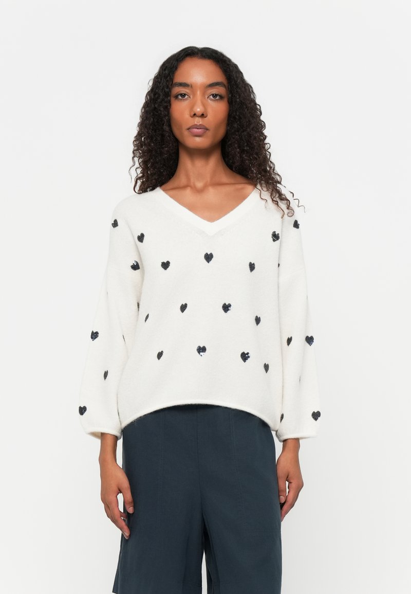 Maglione crema con scollatura a V e maniche oversize, caratterizzato da motivi a cuore neri. Texture morbida con una vestibilità comoda. Abbinato a pantaloni scuri.