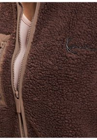 Giacca in pile marrone con una superficie morbida e texturizzata, dotata di una zip beige e di un logo sul davanti, che mette in risalto i suoi colori e materiali a contrasto.