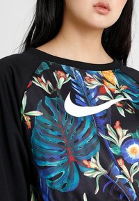 Svart sweatshirt med livfulla tropiska blad- och blommönster, rund halsringning och en framträdande vit Nike-logotyp på framsidan.