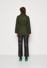 Abrigo de lana verde con cintura ceñida, que presenta un cuello y mangas largas. Usado sobre pantalones de cuero negros y botas negras con detalles en verde.