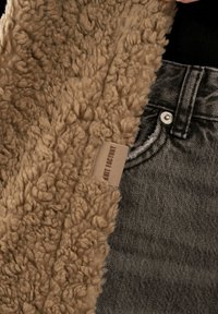 Tan nepleren stof met een fluffy textuur, met een label "Knit Factory." Naast donkergrijze denim met een zichtbare knoop.