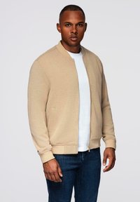 Beżowa kurtka bomberka z zamkiem z przodu, ściągaczami przy mankietach i kieszeniami bocznymi, noszona na białym t-shircie, w połączeniu z niebieskimi dżinsami.