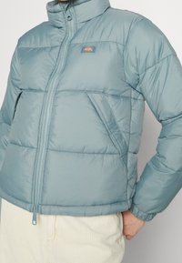 Padded jacket in light blue, featuring a high collar, front zipper, and side pockets with a subtle logo. Smooth texture and compact design.