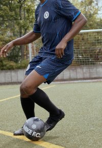 Balón de fútbol negro de Nike con marca blanca sobre césped verde, llevado por un jugador con un uniforme azul con estampado del Chelsea y calcetas negras.