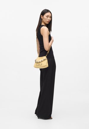 Woman in black sleeveless top and wide-leg pants stands sideways holding a small yellow woven handbag over her shoulder.
