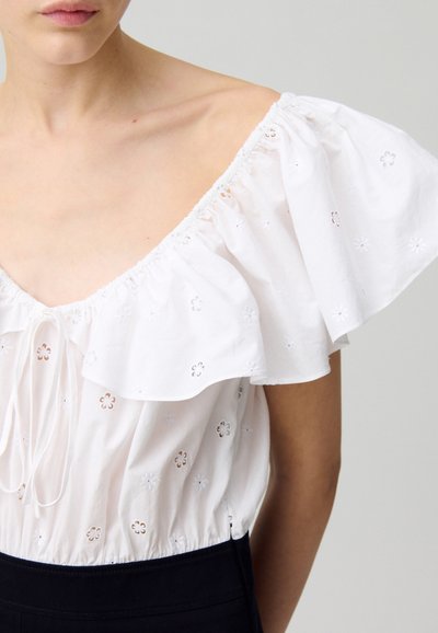 Blouse en coton blanc avec des manches froncées à épaules dénudées et des détails floraux brodés. Dispose d'une taille cintrée et d'une fermeture à lacet.