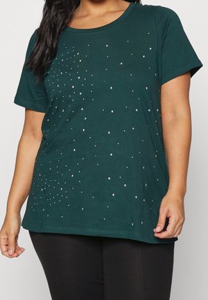 Vrouw draagt een donkergroen topje met korte mouwen en verspreide kleine zilveren studs, gecombineerd met een zwarte broek, zichtbaar van nek tot heupen.