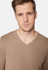 Brązowy sweter w serek wykonany z miękkiego, dzianinowego materiału. Posiada subtelne logo na lewej piersi oraz prążkowane akcenty na dekolcie.