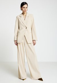 Completo beige su misura con blazer oversize e pantaloni a gamba larga. Il tessuto è liscio con una texture sottile, caratterizzato da una chiusura con un unico bottone.