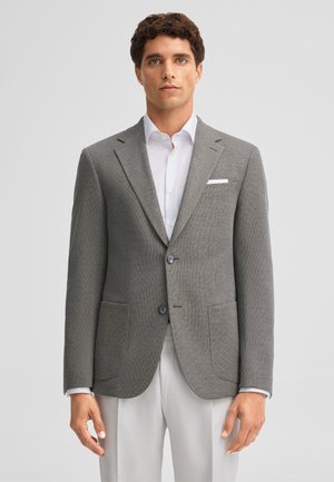 Man wearing grey textured blazer over white dress shirt and light-colored trousers, standing against plain background.