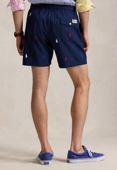 Σορτς σε ναυτικό μπλε με κεντημένα λογότυπα πόλο σε κόκκινο, άσπρο και μπλε. Ελαστική μέση, πίσω τσέπη με κλείσιμο με κουμπί. Φοριούνται με μπλε καμβαδένια παπούτσια.