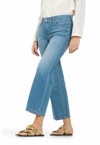 Žena v světle modrých zkrácených rozšířených džínách, bílém dlouhém rukávu a zlatých pletených plochých sandálech, stojící s překříženýma nohama.