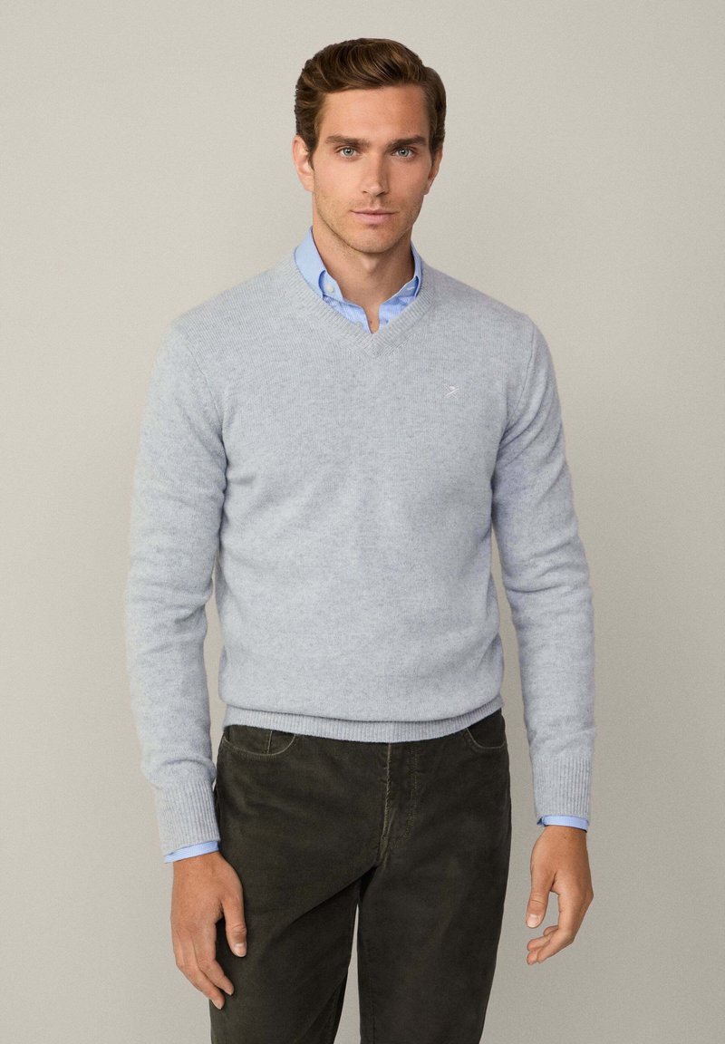 Hellgrauer gestrickter Pullover mit V-Ausschnitt und gerippten Bündchen, über einem hellblauen Kragenhemd getragen, kombiniert mit dunklen Hosen.