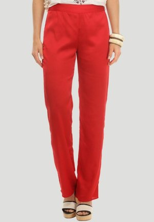 Mujer con pantalones rojos brillantes de pierna recta, sandalias blancas de plataforma y una camiseta blanca con pulseras en la muñeca derecha.