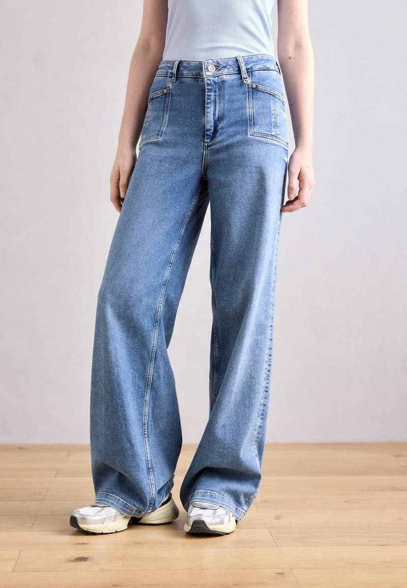 Personne portant un jean large bleu clair et des baskets blanches, debout sur un sol en bois devant un fond uni.