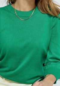 Grüner Strickpullover mit Rundhalsausschnitt und gerippten Bündchen. Der Stoff wirkt weich und glatt und hat minimalistische Details. Eine schlichte goldene Halskette ist sichtbar.