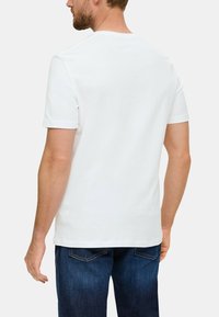 Camiseta de algodón blanca con escote redondo y mangas cortas, mostrada desde atrás. Llevada con jeans de mezclilla azul oscuro.