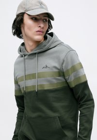 Młoda osoba ubrana w zielono-szaro-blokowany hoodie i szary czapkę baseballową, patrząca w bok na jednolitym tle.
